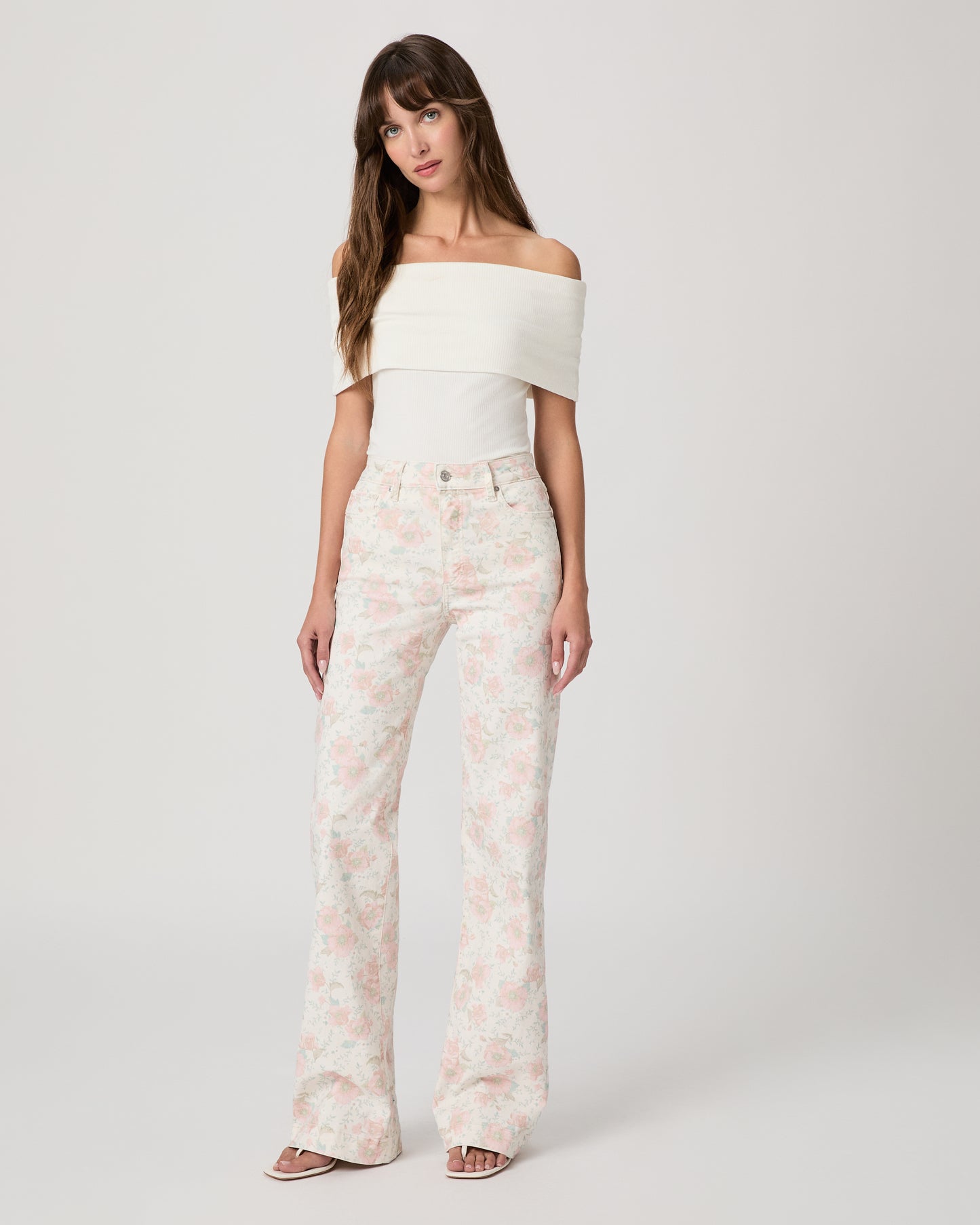 Woman wearing a white top and flower printed jeans paired with white sandals on a white background