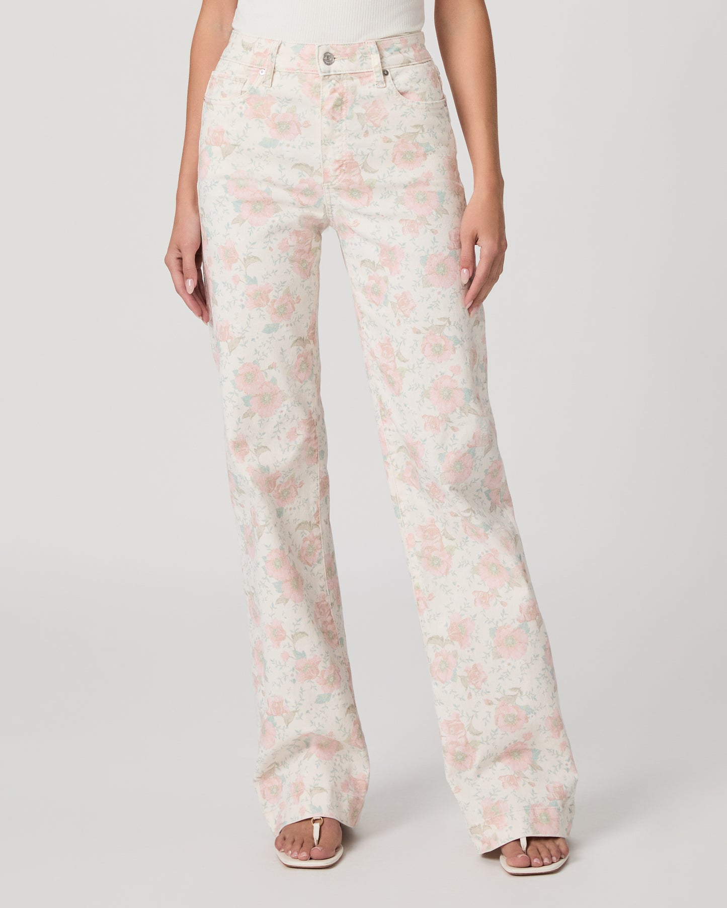 Woman wearing a white top and flower printed jeans paired with white sandals on a white background