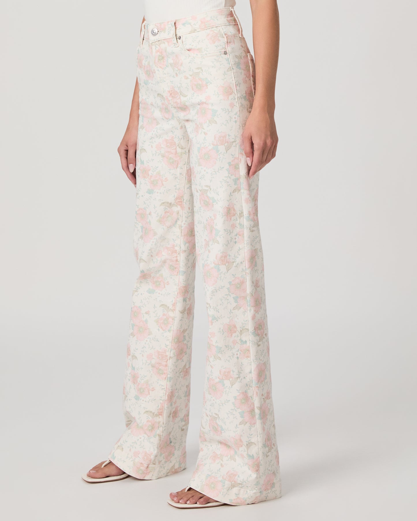 Woman wearing a white top and flower printed jeans paired with white sandals on a white background