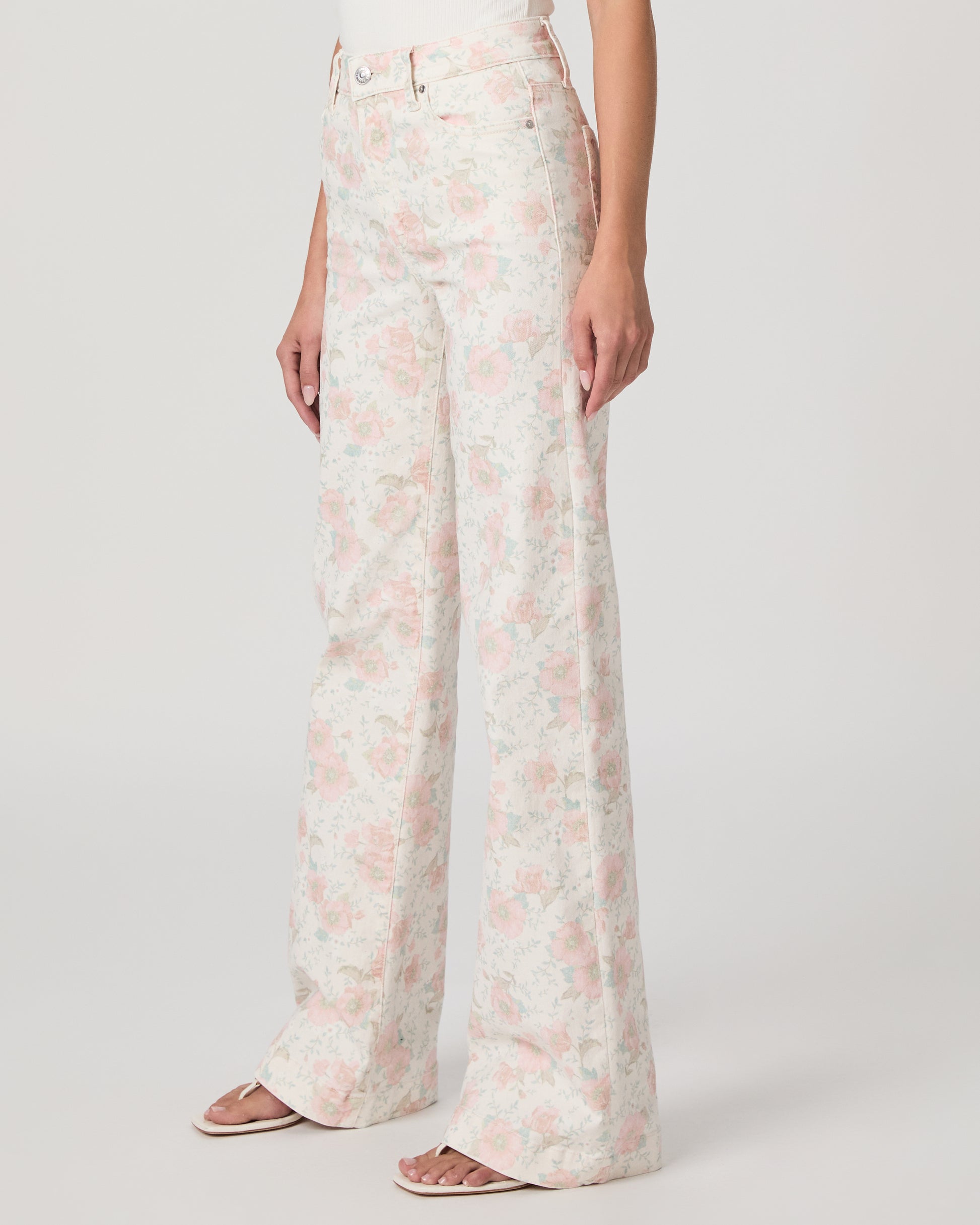 Woman wearing a white top and flower printed jeans paired with white sandals on a white background