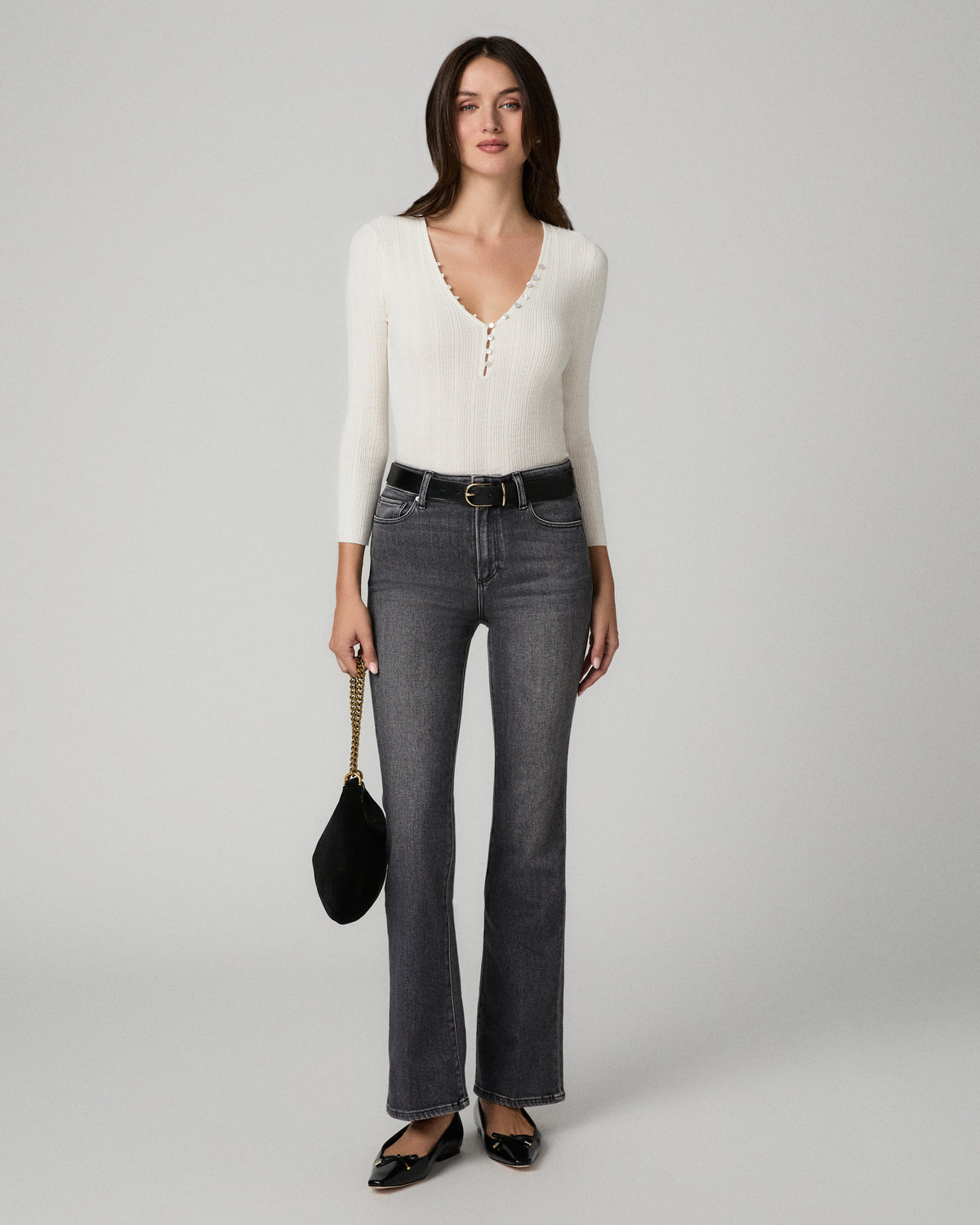 A woman stands against a light gray studio background wearing a fitted off-white ribbed knit long-sleeve top with a scalloped V-neckline, dark gray wide-leg jeans, a black belt, black flats, and holding a small black handbag.