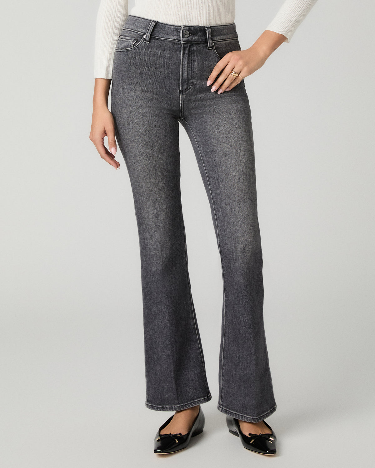 A model wearing a pair of dark gray high-rise flare jeans, styled with a white ribbed long-sleeve top and black patent leather flats, against a light gray studio background.