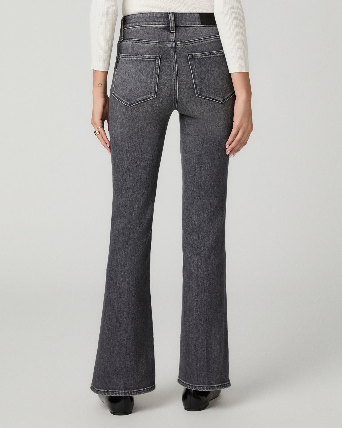 Rear view of a model wearing dark gray high-rise flare jeans with a white long-sleeve top and black shoes, set against a light gray studio background.