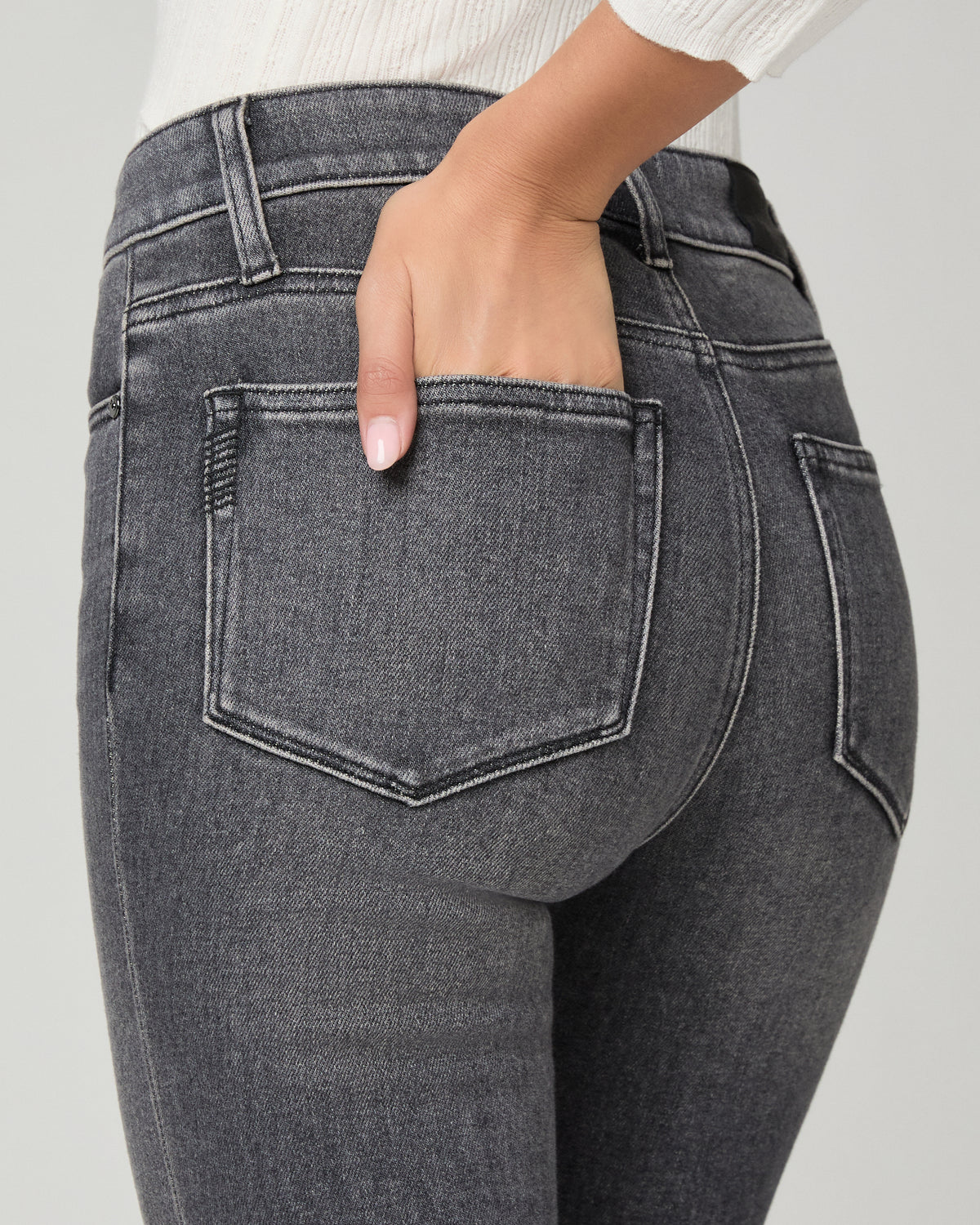 Close-up back view of a person wearing dark gray denim jeans, with one hand tucked into the back pocket against a light gray studio background.