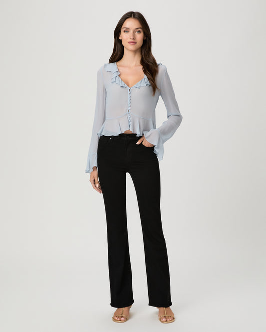 A woman modeling a light blue sheer long-sleeve blouse with ruffle trim and a button front, paired with black pants, against a plain light gray studio background.