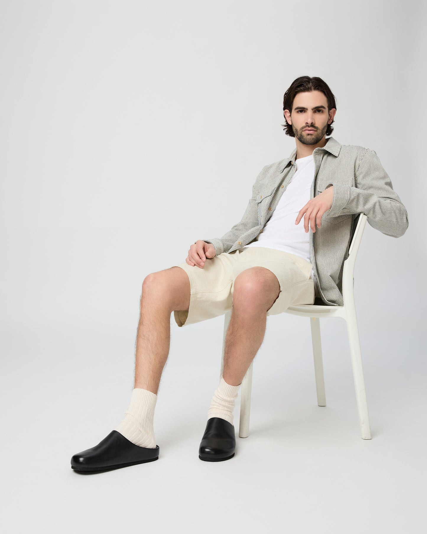 Man sitting on a chair wearing a light gray jacket, white shirt, beige shorts, and black shoes on a white background