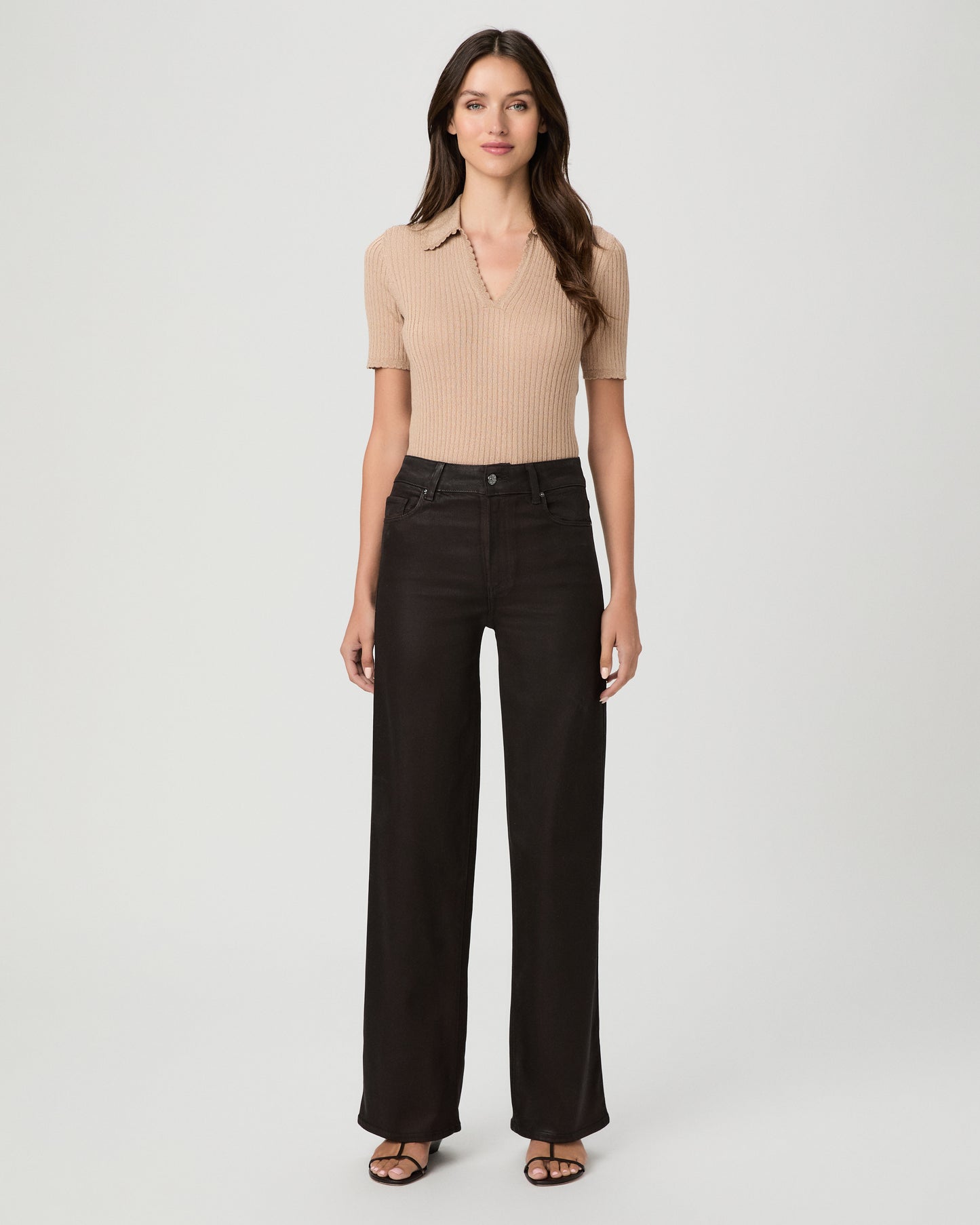 A woman modeling a pair of dark burgundy pants with cream color top and brown sandals against a light gray studio background.