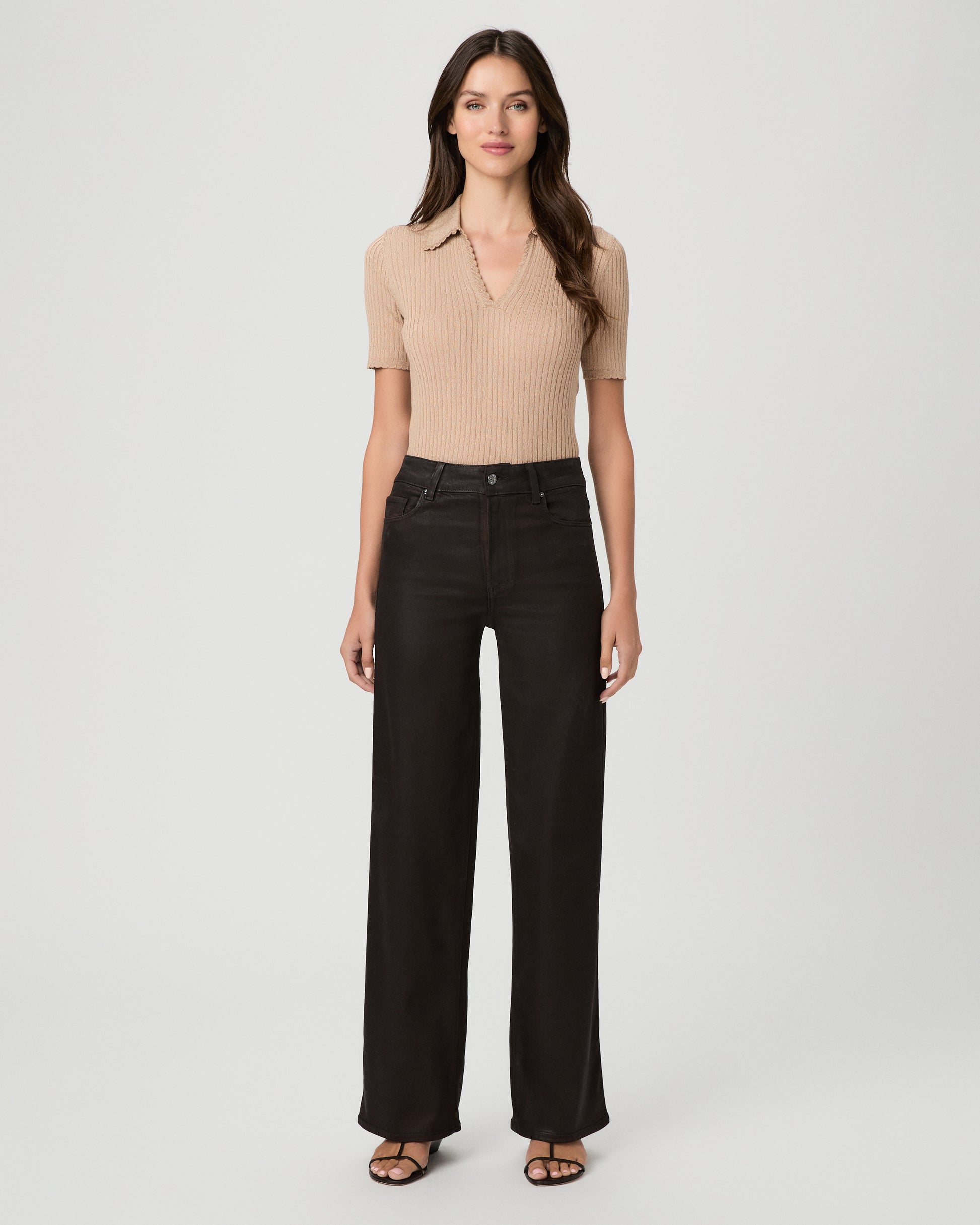 A woman modeling a pair of dark burgundy pants with cream color top and brown sandals against a light gray studio background.