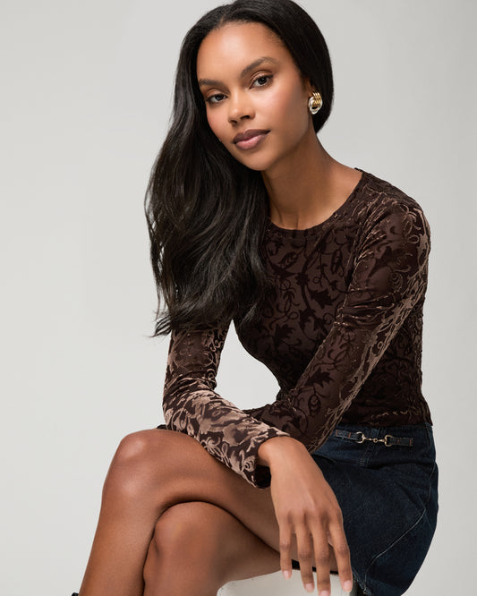 Seated close-up of a woman wearing a brown velvet burnout long-sleeve top with a floral pattern, one hand resting near her knee, paired with a dark denim mini skirt.
