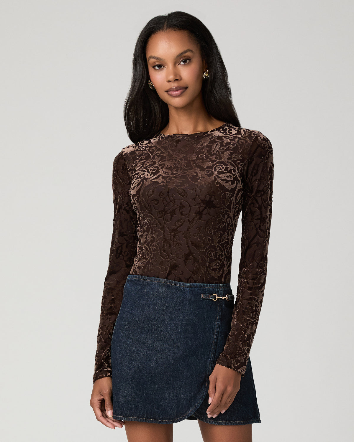 Close-up front view of a woman wearing a brown velvet burnout long-sleeve crew-neck top with a floral pattern, over a dark denim mini skirt.