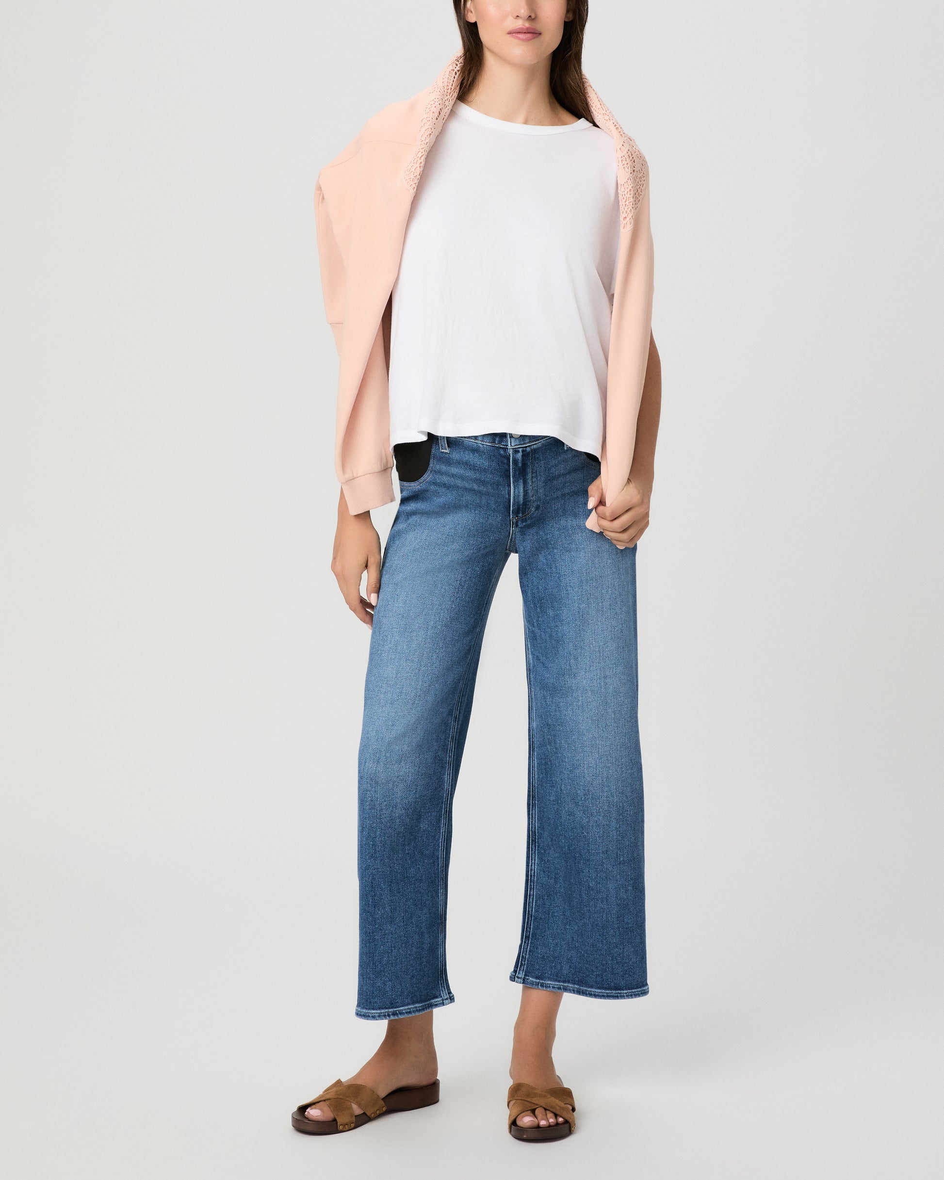 A model wearing a white crewneck t-shirt, blue wide-leg cropped jeans, a light pink open-front cardigan, and brown slide sandals, photographed against a neutral light gray studio background.