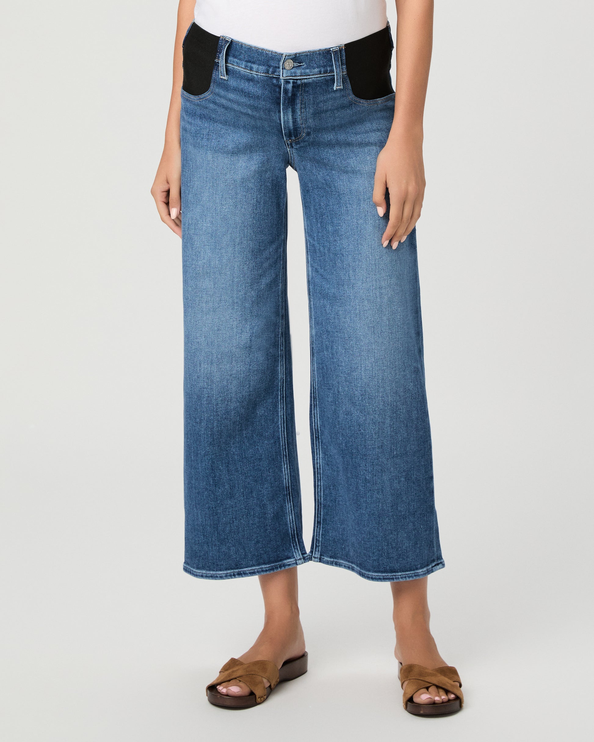 A model wearing a white crewneck t-shirt, blue wide-leg cropped jeans, a light pink open-front cardigan, and brown slide sandals, photographed against a neutral light gray studio background.