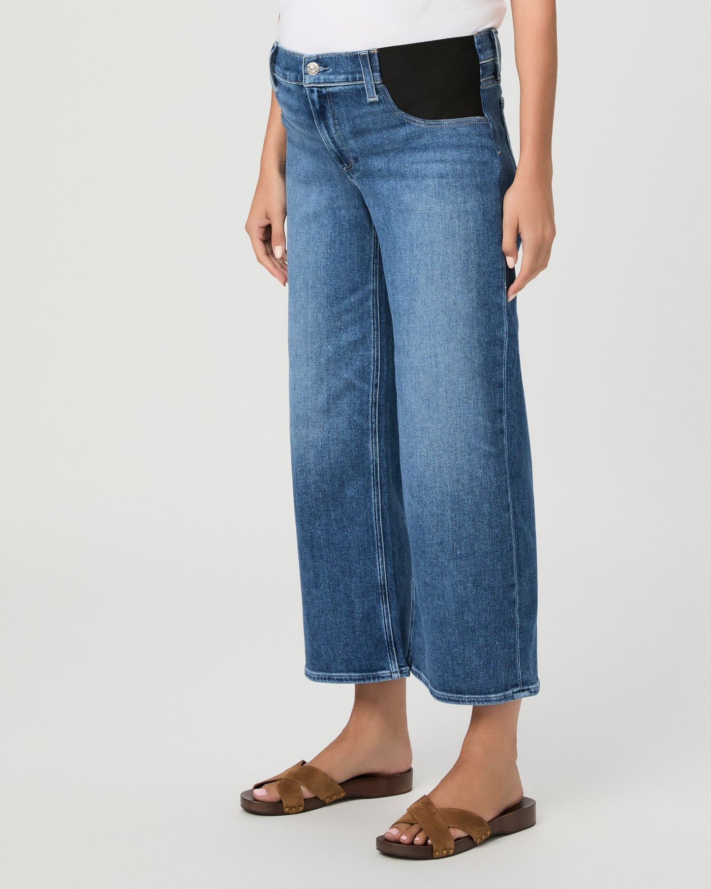 A model wearing a white crewneck t-shirt, blue wide-leg cropped jeans, a light pink open-front cardigan, and brown slide sandals, photographed against a neutral light gray studio background.