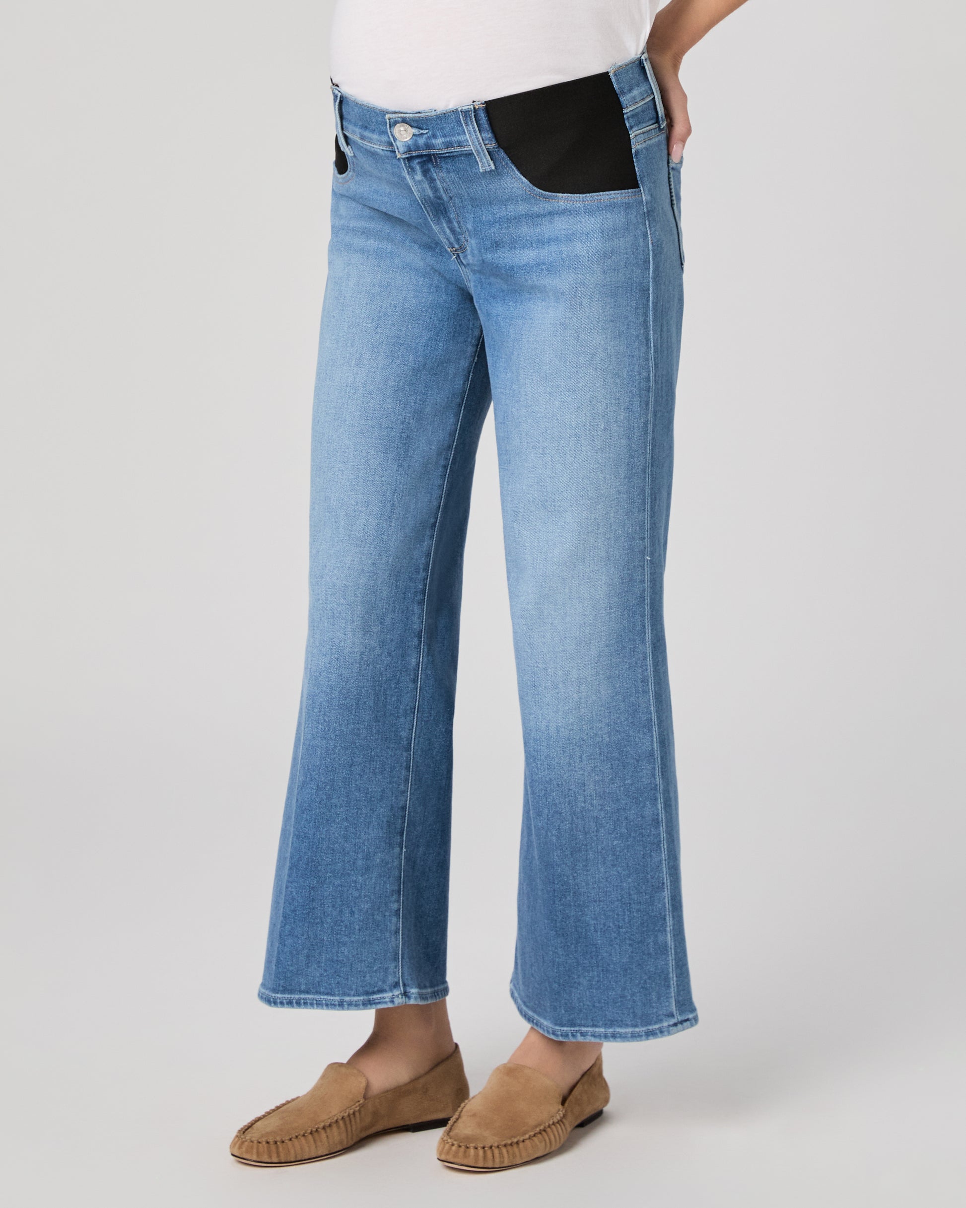 Woman wearing a maternity blue jeans and a white top paired with brown flats on a plain background