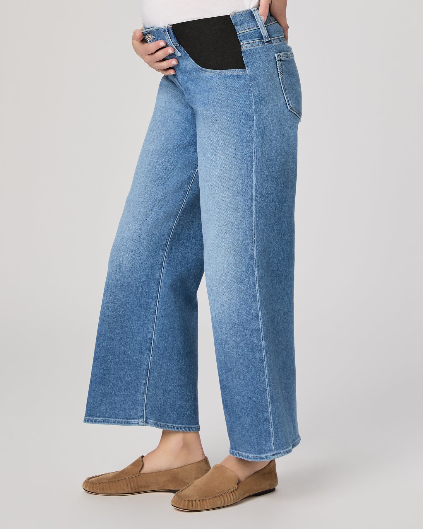 Woman wearing a maternity blue jeans and a white top paired with brown flats on a plain background