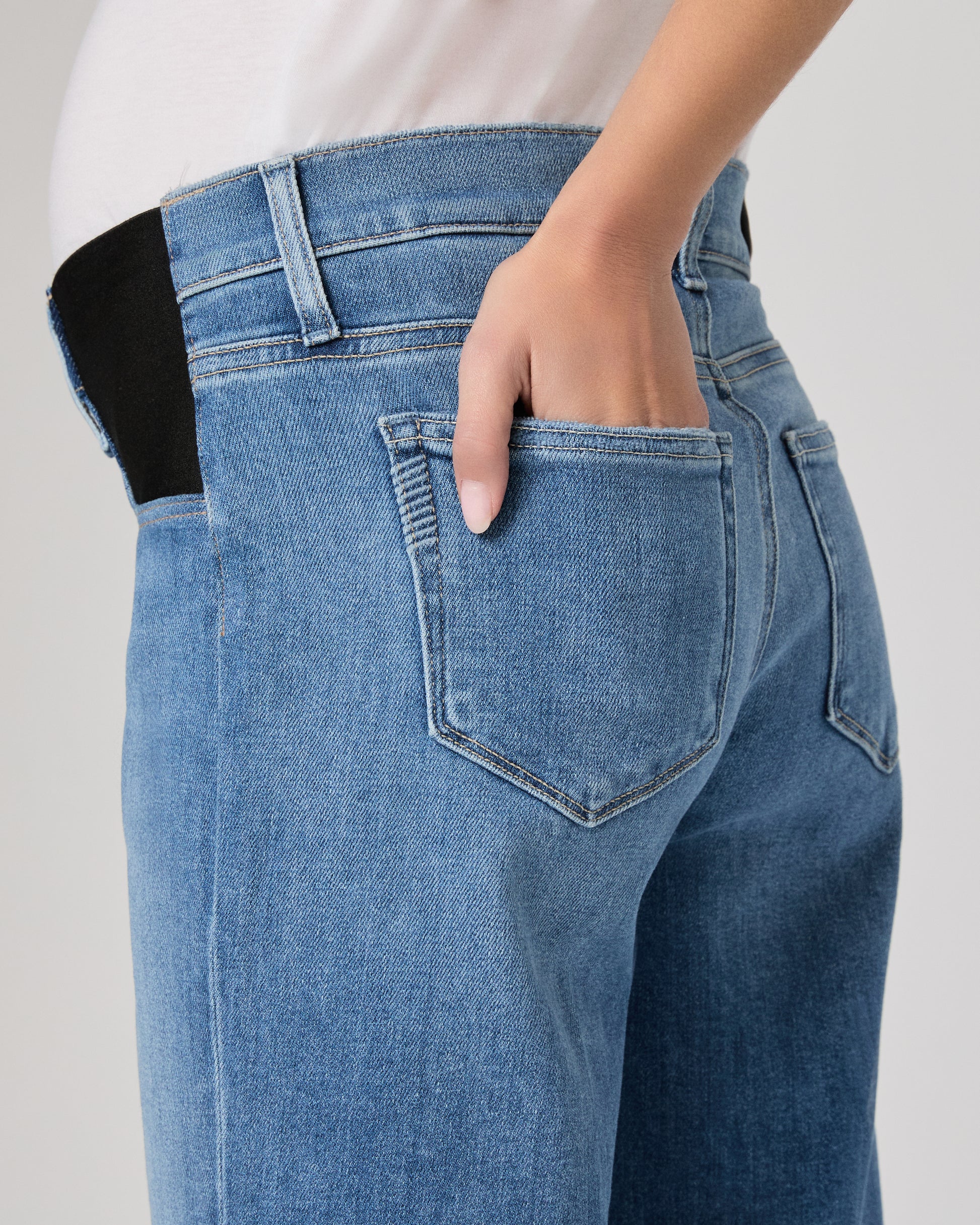 Woman wearing a maternity blue jeans and a white top paired with brown flats on a plain background