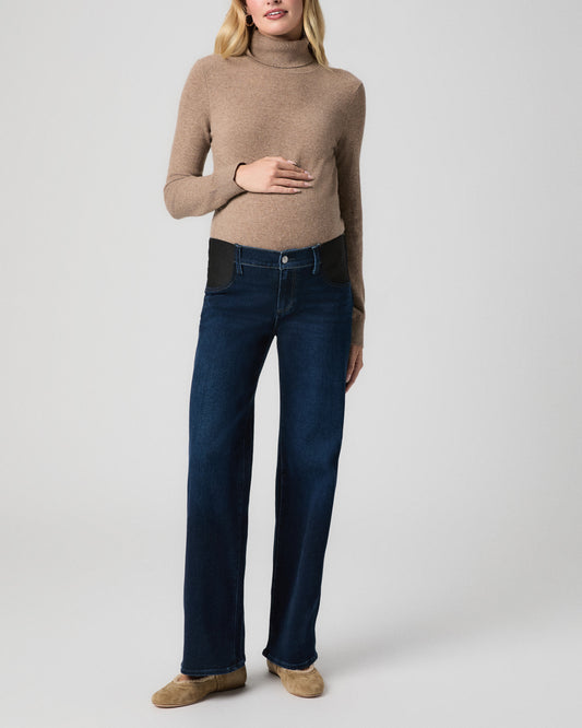 Pregnant model standing facing forward, one hand on her belly, wearing a camel turtleneck sweater, Anessa Maternity Jeans in Amagansett dark wash with black side panels, and tan flats