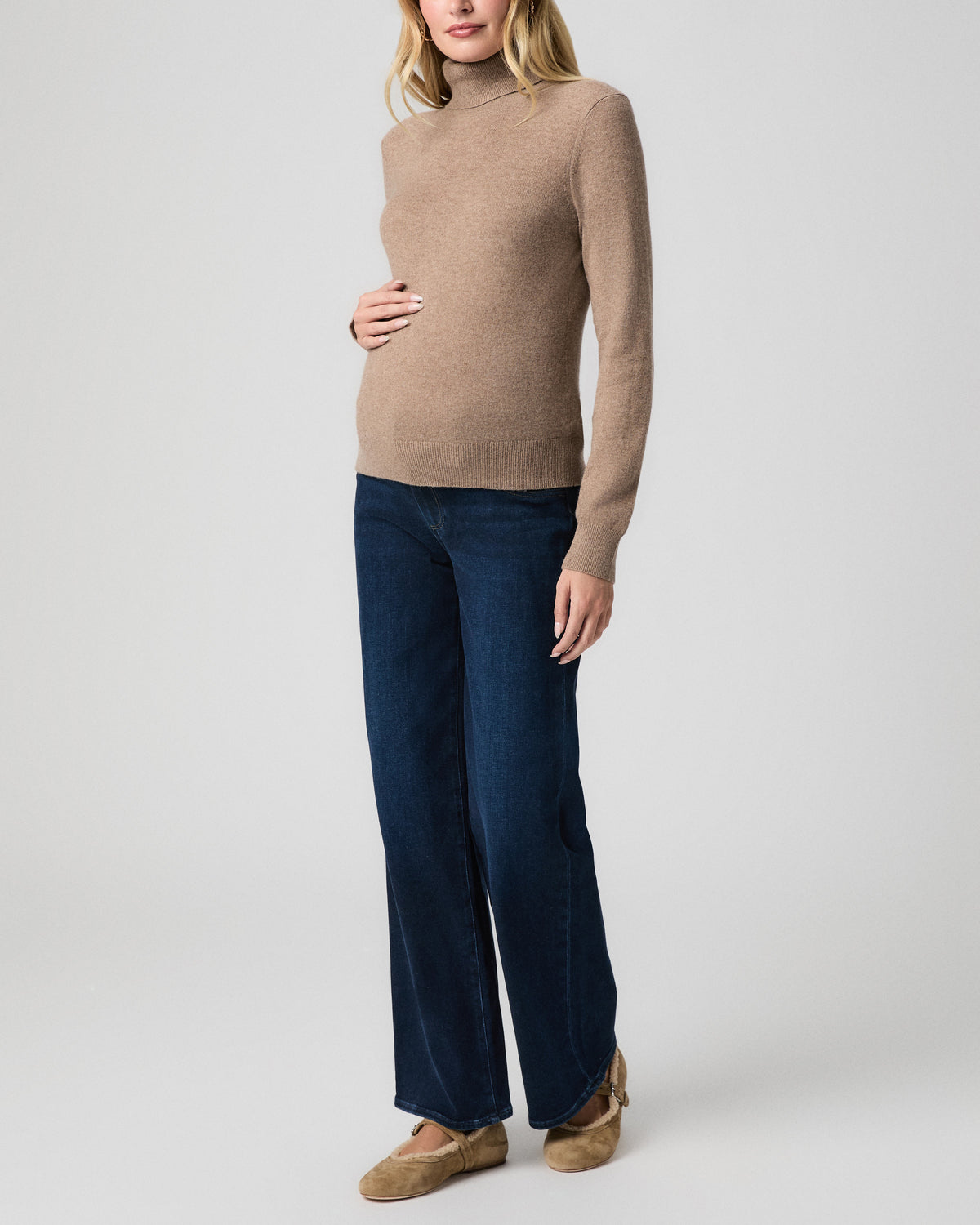 Pregnant model standing at a three-quarter angle, one hand on her belly, wearing a camel turtleneck sweater, Anessa Maternity Jeans in Amagansett, and tan flats