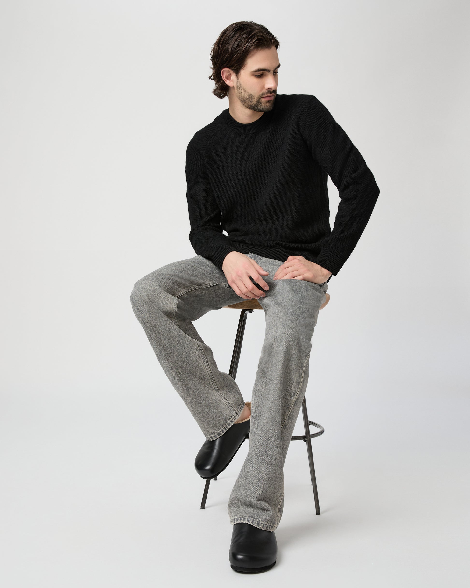 Man sitting on a stool wearing a black sweater and gray jeans against a white background