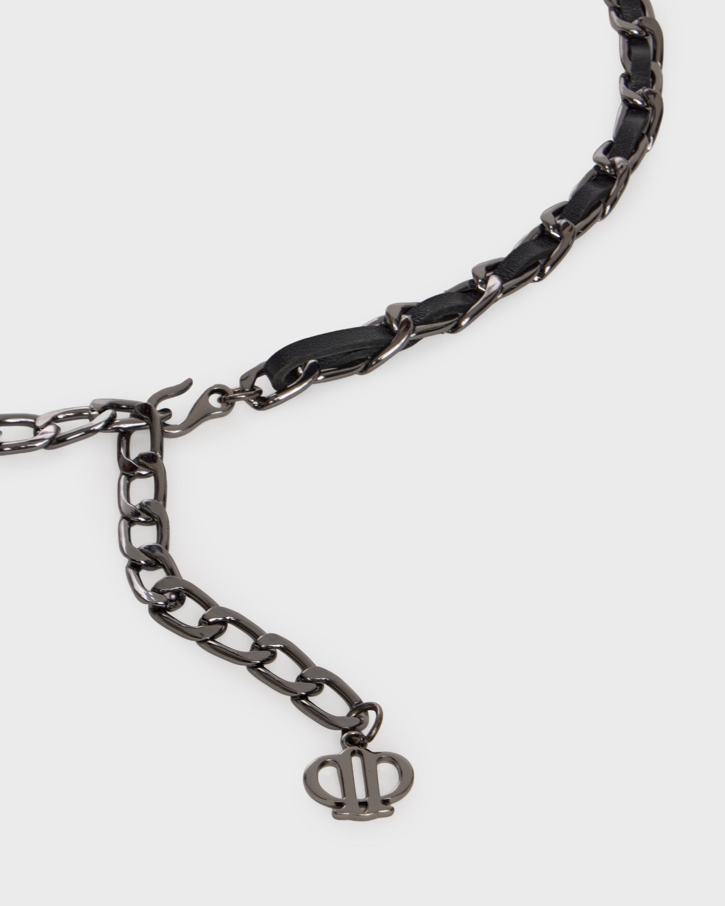 A close-up of a dark metallic chain necklace featuring a black leather accent segment and a small interlocking monogram charm, shown on a light gray background.
