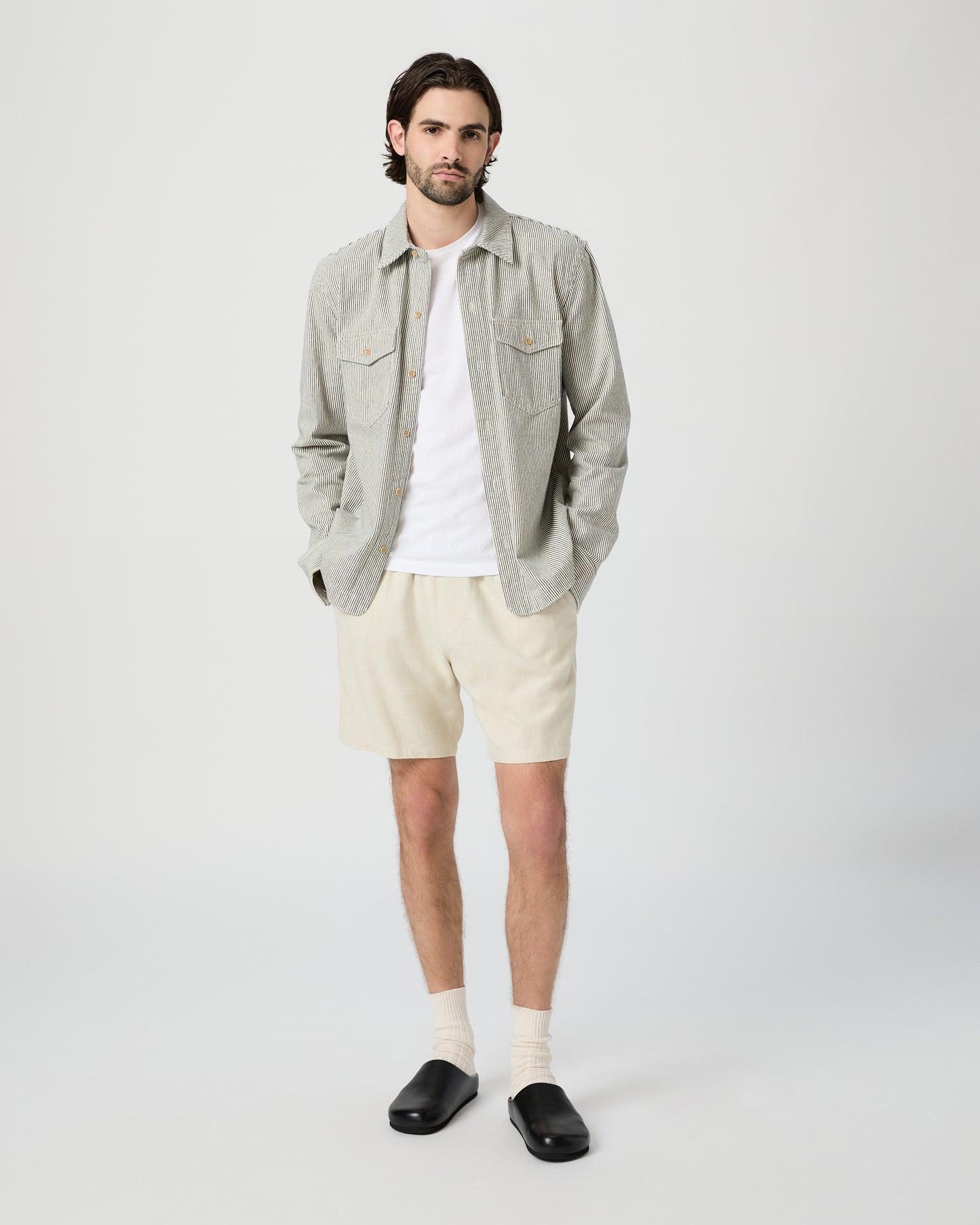 Man wearing a light gray shirt, white t-shirt, beige shorts, and black shoes on a white background