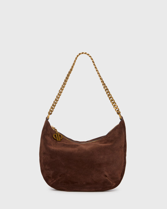 A dark brown suede crescent shoulder bag with a gold-tone chain strap and a small gold-tone metal logo charm, shown against a light gray background.