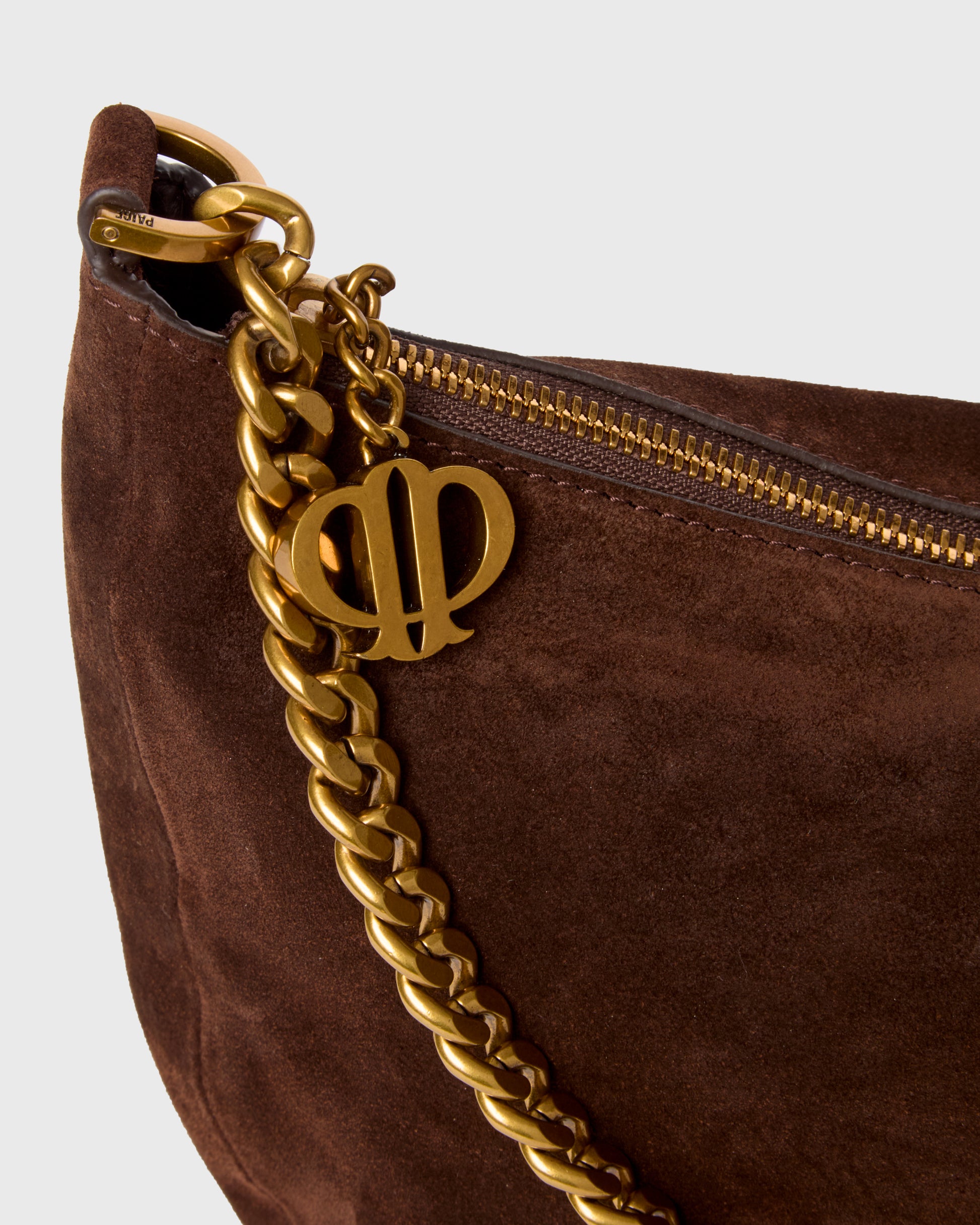 Close-up of a dark brown suede shoulder bag with a chunky gold chain strap and a gold interlocking letters charm on a light gray background.