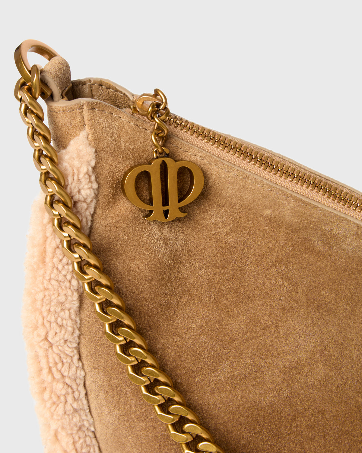 Close-up of a taupe suede shoulder bag with a cream sherpa lining, a gold chain strap, and a gold metal monogram charm on a light gray background.