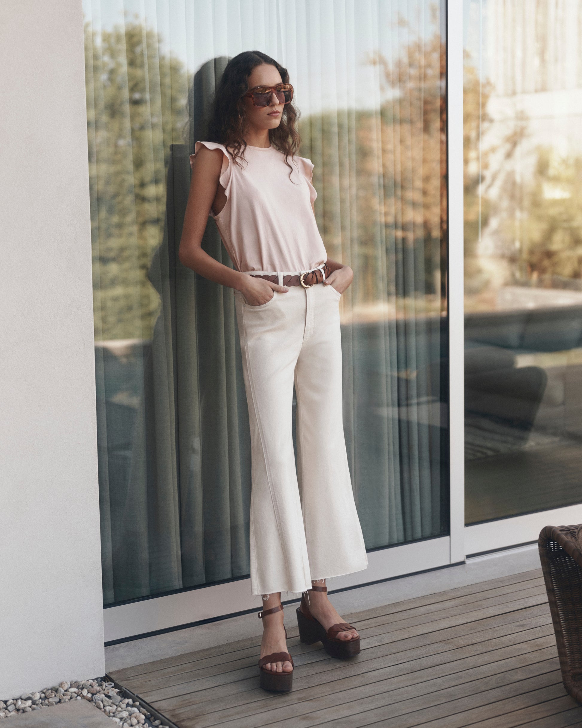 Woman in a pink top and white pants standing on a wooden deck with large glass doors in the background.