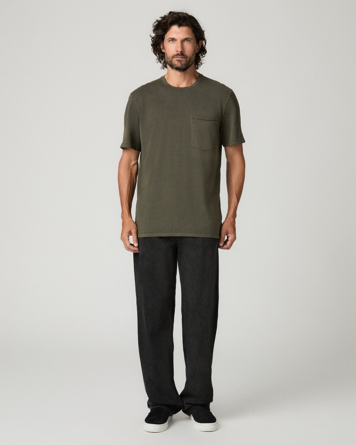 Full-length front view of a man standing casually in a vintage fatigue green crew-neck tee with a chest pocket, paired with black straight-leg pants and black sneakers.