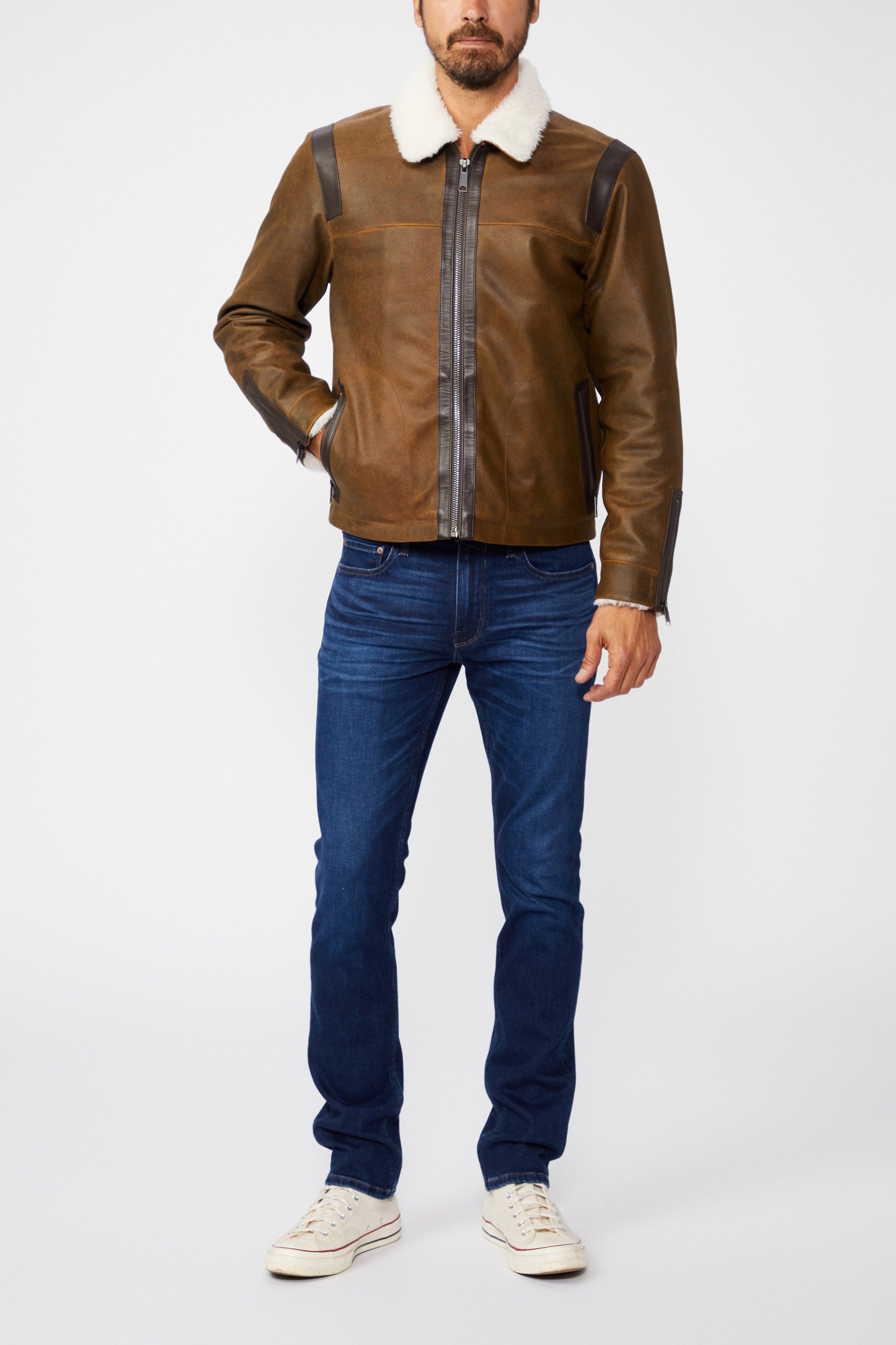 Man wearing a brown leather jacket with a white collar, blue jeans, and white sneakers on a white background