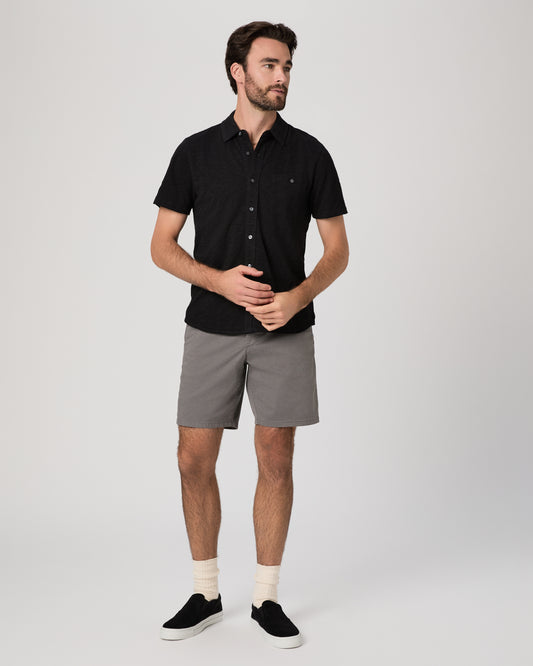 Man wearing black short sleeve shirt paired with grey shorts and black sneakers against a white background