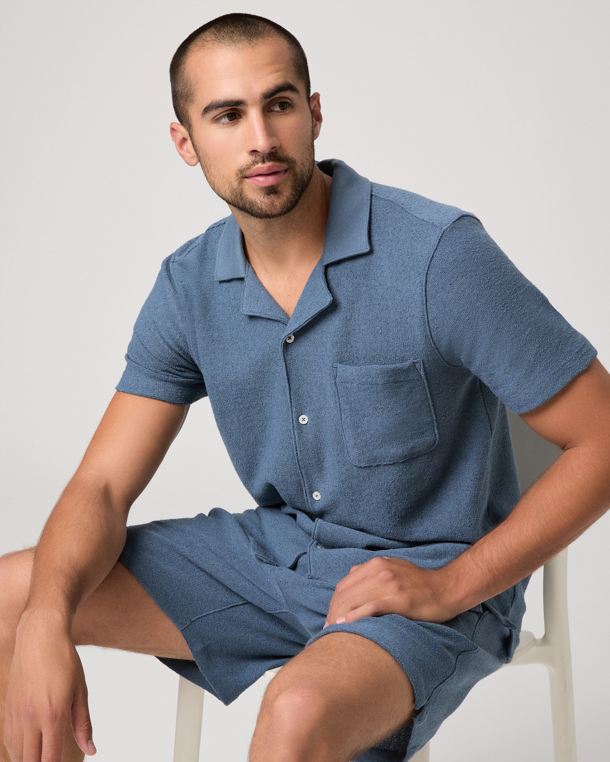 Man seating on a chair wearing a blue short-sleeve shirt and shorts on a light gray background