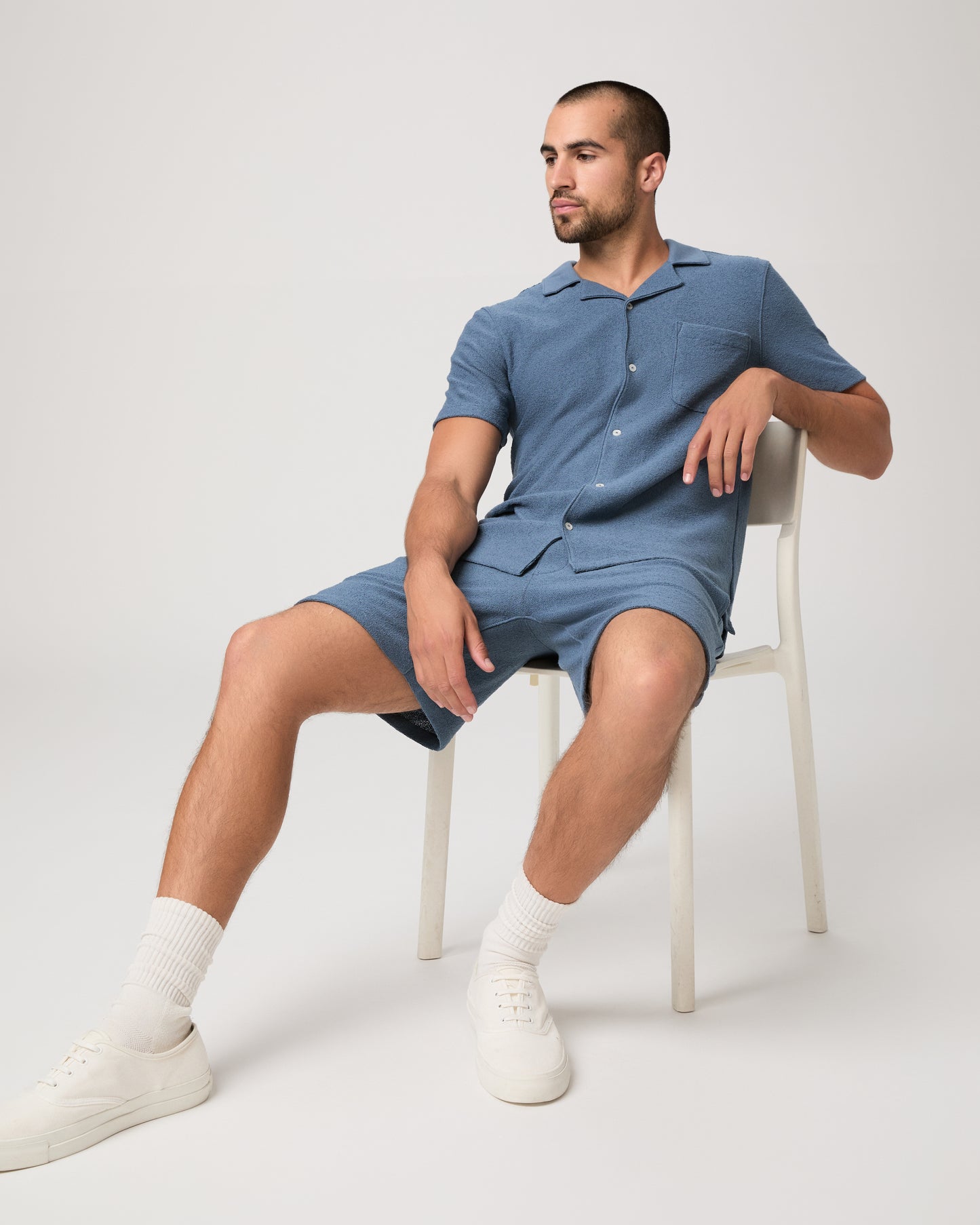 Man seating on a chair wearing a blue short-sleeve shirt and shorts on a light gray background