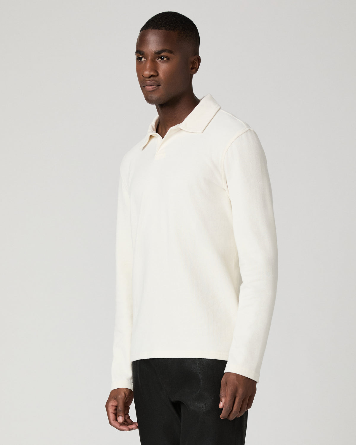 Three-quarter front view of a man wearing the natural off-white long-sleeve polo shirt, showing the ribbed knit texture and clean placket, paired with black trousers.