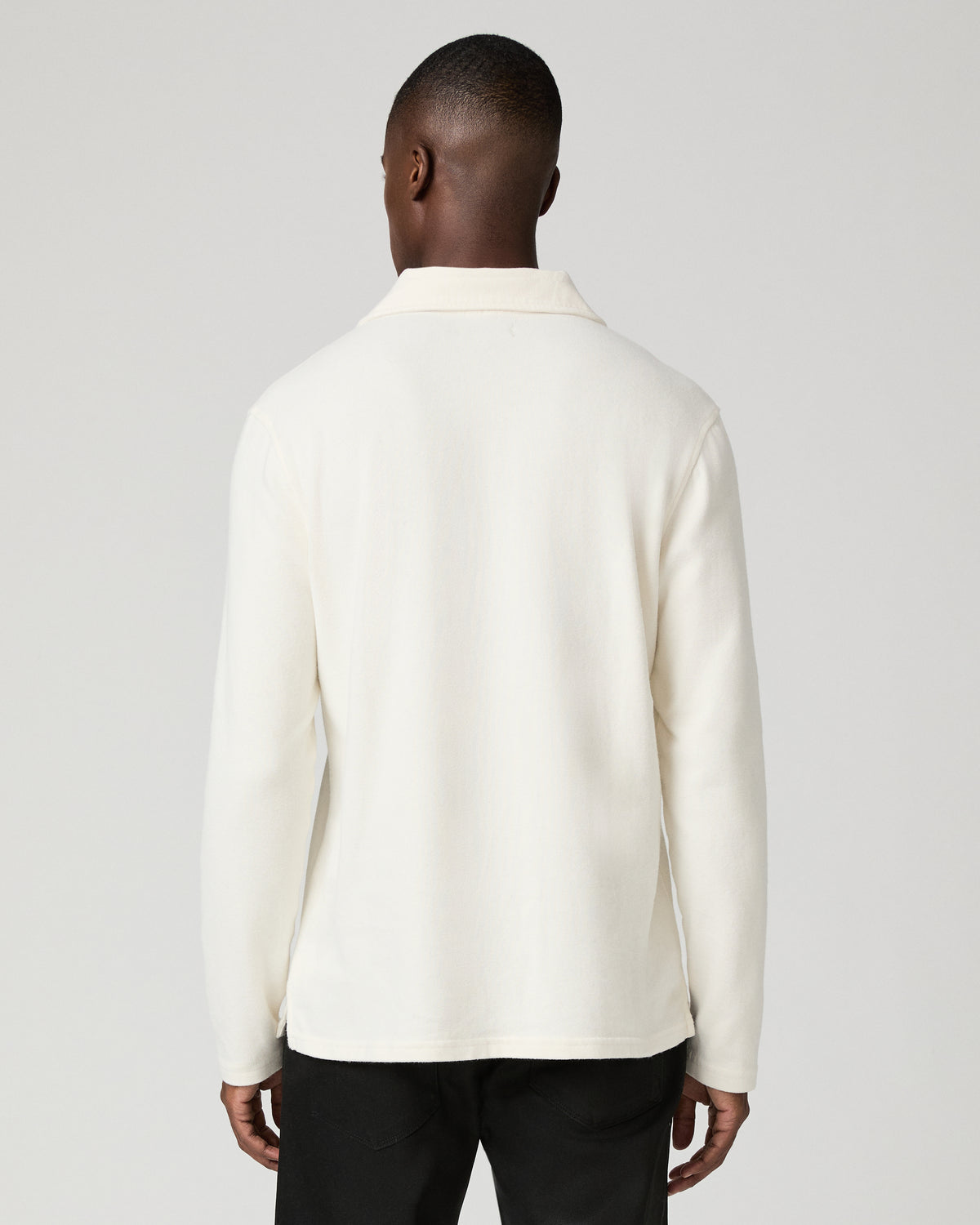 Back view of a man wearing the natural off-white long-sleeve polo shirt, highlighting the ribbed knit fabric and straight hem, paired with black trousers.