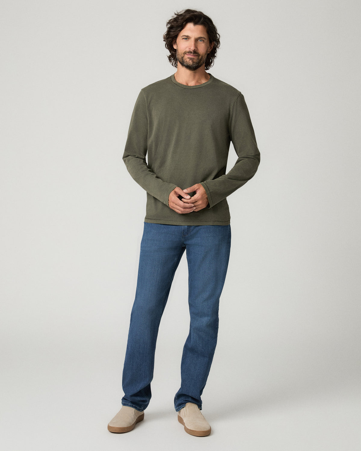 Full-length front view of a man standing with hands clasped, wearing the vintage fatigue green Enzo Shirt, a long-sleeve crewneck knit top with raw-edge cuffs, paired with straight-leg blue jeans and tan suede loafers.