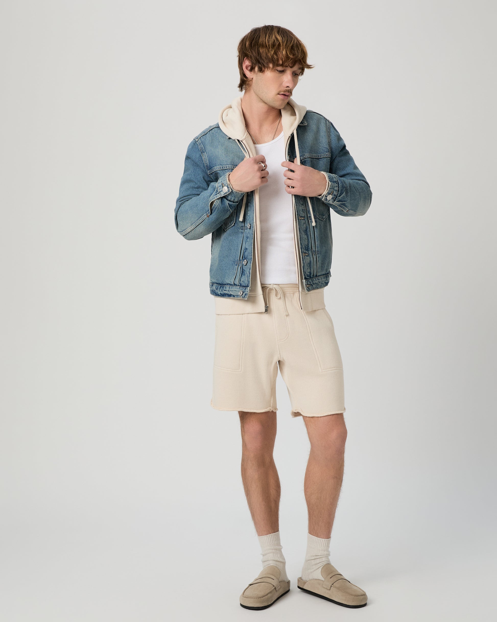 Man wearing a denim jacket, white shirt, beige shorts, and shoes on a plain background