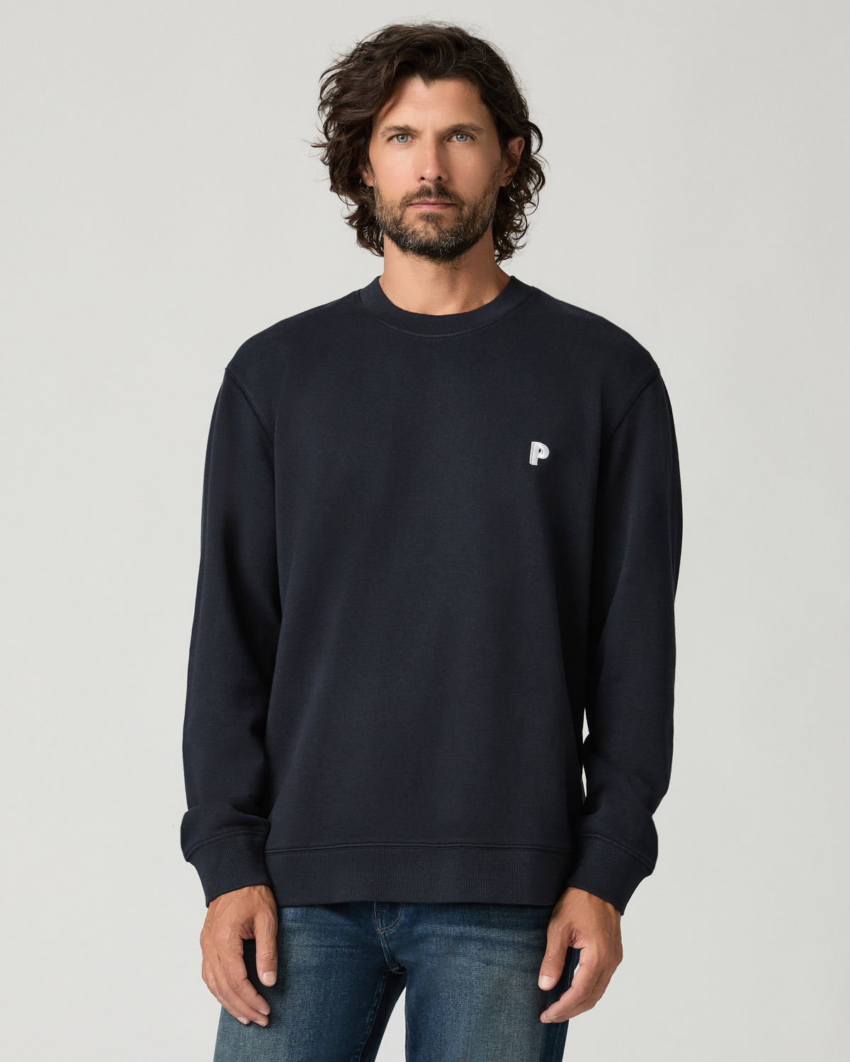 Cropped front view of a man with curly hair wearing a deep navy crewneck pullover with a small white embroidered logo on the left chest.