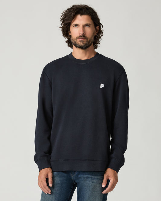 Cropped front view of a man with curly hair wearing a deep navy crewneck pullover with a small white embroidered logo on the left chest.