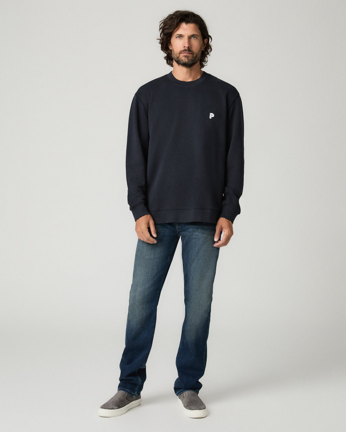 Full-length front view of a man standing in a deep navy crewneck pullover with a small white chest logo, paired with dark wash straight-leg jeans and gray suede slip-on sneakers.