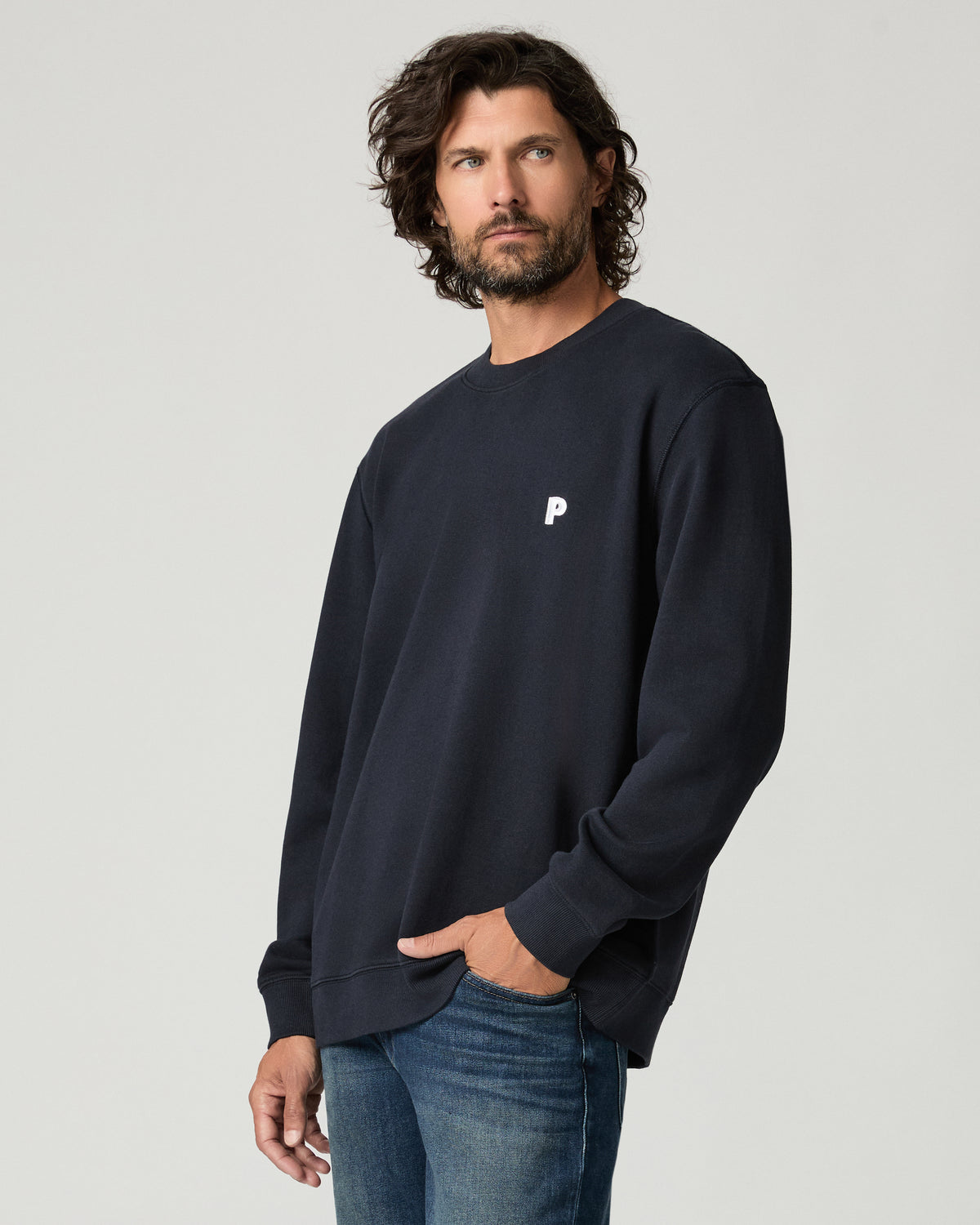 Three-quarter side view of a man with one hand in his jeans pocket, wearing a deep navy crewneck pullover with a small white chest logo.
