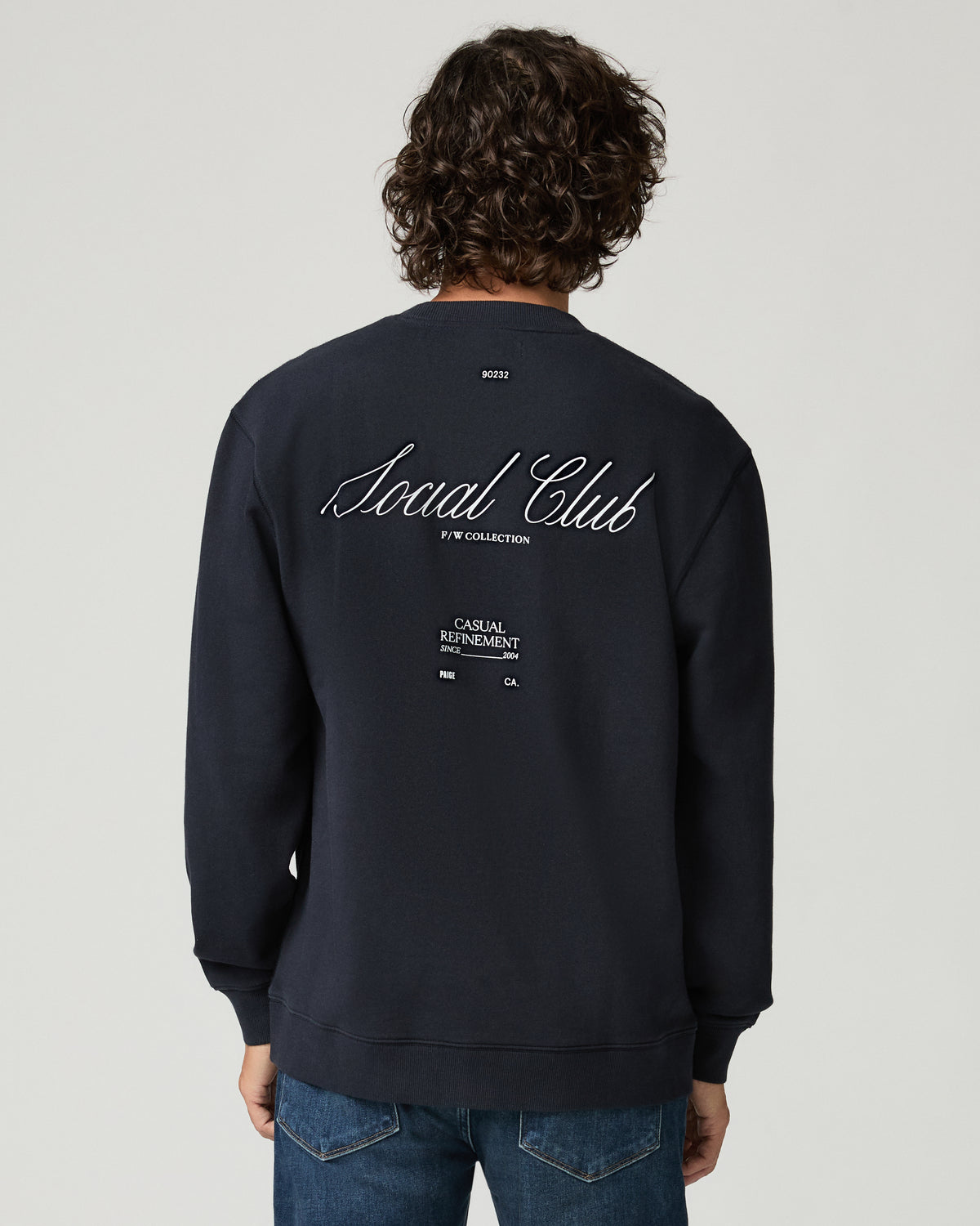 Back view of a man wearing a deep navy pullover featuring a large 'Social Club' script graphic print across the upper back with smaller text beneath it.