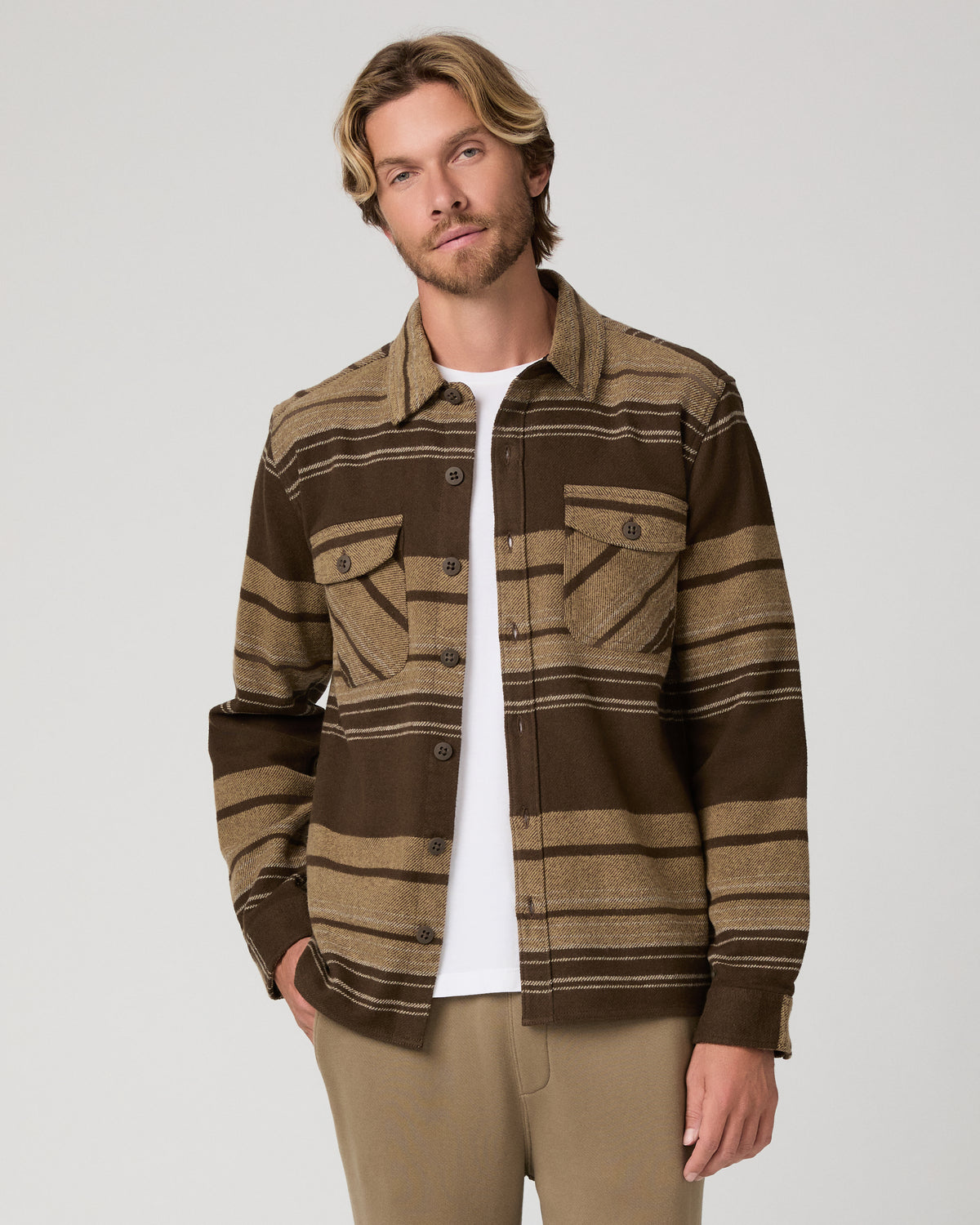 A man wearing a brown and tan striped button-up shirt jacket over a white t-shirt and khaki pants, photographed against a light gray studio background.