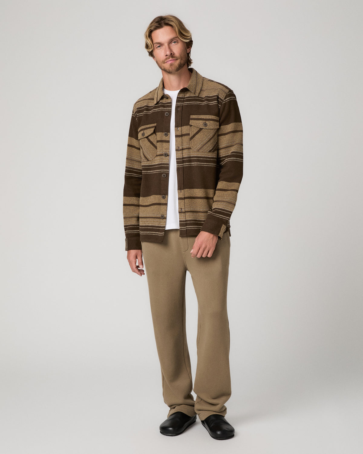 A man wearing a brown and tan striped button-up shirt jacket over a white t-shirt and khaki pants, black shoes photographed against a light gray studio background.