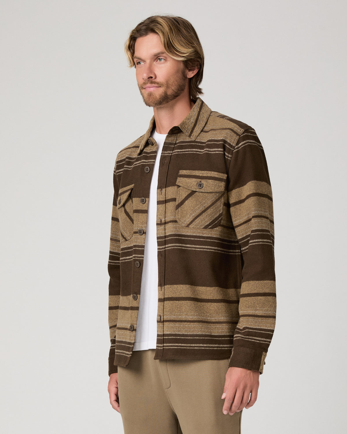A man wearing a brown and tan striped button-up shirt jacket over a white t-shirt and khaki pants, photographed against a light gray studio background.