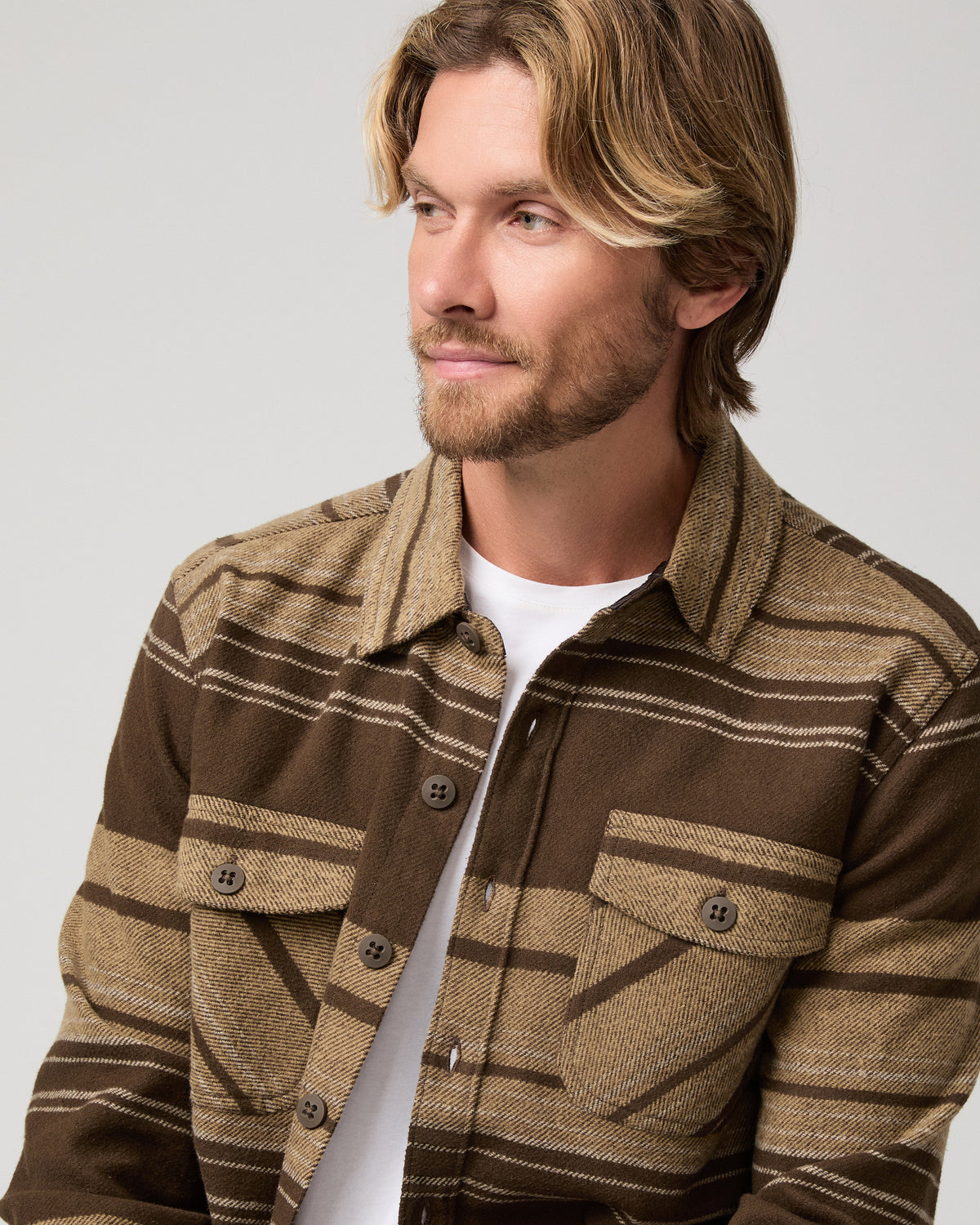A man wearing a brown and tan striped button-up shirt jacket over a white t-shirt and khaki pants, photographed against a light gray studio background.
