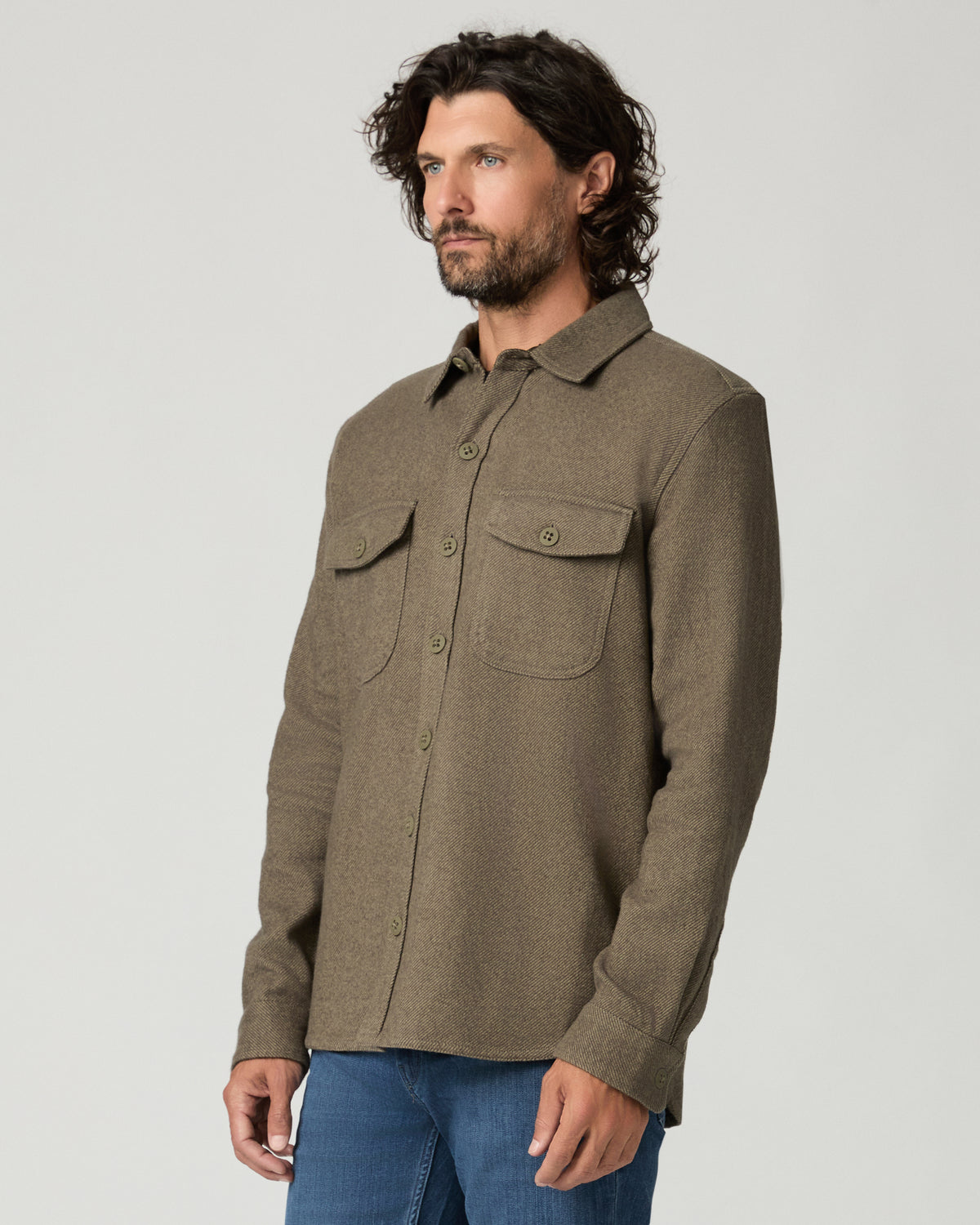 Angled three-quarter front view of the buttoned moss green overshirt, showing the flap chest pocket and button placket, hand in pocket.