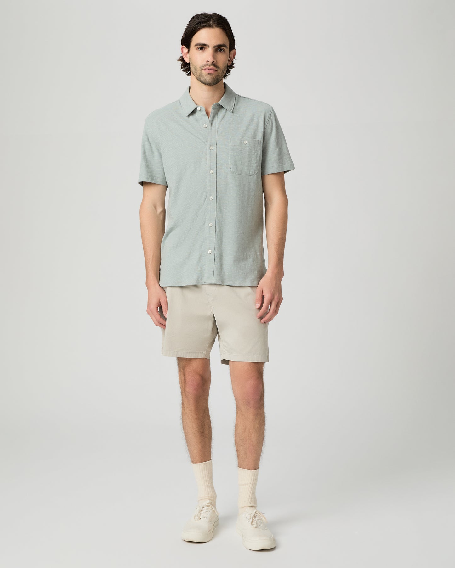 Man wearing a light green button-up shirt and beige shorts on a white background