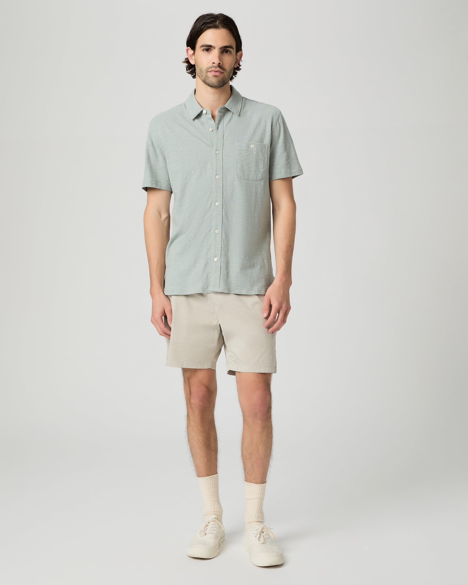 Man wearing a light green button-up shirt and beige shorts on a white background